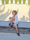Niño con auriculares y el uso de teléfonos inteligentes mientras está sentado en el monopatín en el parque de skate urbano soleado - foto de stock