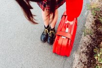 Mulher em botas elegantes com mala vermelha de pé na estrada — Fotografia de Stock