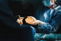 Médico em vestido cirúrgico segurando preparado implante de silicone para mamoplastia e enfermeira de colheita auxiliando durante a operação — Fotografia de Stock