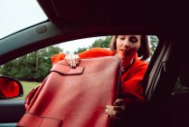 Женщина кладет винтажный красный чемодан на переднее сиденье автомобиля через окно — стоковое фото
