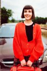 Очаровательная привлекательная женщина со стильными волосами в красном свитере с красным чемоданом ждет поездки в машине позади на дороге — стоковое фото