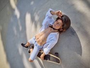 Blick von oben auf ein Kind im Helm, das mit geschlossenen Augen auf dem Boden im Skatepark mit Schatten liegt — Stockfoto