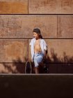 Garçon en chemise blanche debout appuyé sur le mur avec planche à roulettes dans la lumière du soleil regardant loin — Photo de stock