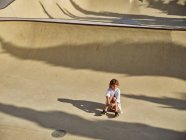 Вид зверху на дитину в шоломі, що сидить і охолоджується на землі в скейтпарку з тінями, дивлячись далеко — стокове фото