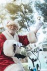 Homem idoso em traje de Papai Noel sentado em ciclo, tocando sino e olhando para longe — Fotografia de Stock