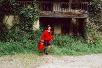Jovem mulher em vermelho com grande mala vermelha correndo no campo contra casa abandonada — Fotografia de Stock