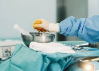 Recadrage main du médecin en robe bleue et gant blanc serrant tampon plein d'iode dans un bol pendant la chirurgie — Photo de stock