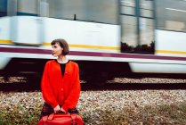 Femme élégante en pull rouge avec valise rouge regardant le long tout en train rapide chevauchant sur le chemin de fer derrière le dos — Photo de stock