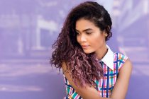 Портрет чарівної молодої етнічної молодої жінки з кучерявим волоссям, що нахиляє голову і дивиться на фіолетову скляну стіну — стокове фото