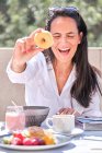 Frau sitzt am Tisch mit serviertem Frühstück auf offener sonnenbeschienener Terrasse, während sie Donut hält und vorführt und mit geschlossenen Augen auf verschwommenem Hintergrund lacht — Stockfoto