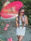 Schlanke junge Frau im Sommeroutfit mit Regenschirm trinkt Getränk in der Nähe blühender Bäume — Stockfoto