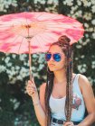 Magro jovem mulher em roupa de verão e óculos de sol com guarda-chuva de pé perto de árvores florescendo — Fotografia de Stock
