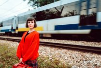 Стильная женщина в красном свитере с красным чемоданом, смотрящая вперед, в то время как скоростной поезд едет по железной дороге за спиной — стоковое фото