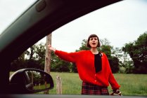 Молодая женщина в красном свитере ловит машину на дороге в сельской местности — стоковое фото
