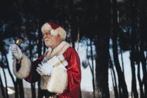 Homme âgé joyeux en costume du Père Noël debout avec présent et cloche dans les mains gantées regardant loin sur fond de nature — Photo de stock