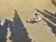 View from above of child in helmet sitting and riding skateboard with shadows — Stock Photo
