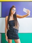 Симпатичная девушка-подросток со стильными дредами, делающими селфи на смартфоне в красочной комнате — стоковое фото