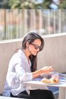 Hübsche freundliche Dame mit Sonnenbrille sitzt am gedeckten Tisch auf der offenen sonnenbeschienenen Terrasse und isst rosa Joghurt, während sie auf ihren Partner wartet — Stockfoto
