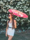 Magro jovem mulher em roupa de verão e óculos de sol com guarda-chuva de pé perto de árvores florescendo — Fotografia de Stock