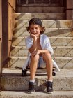 Garçon joyeux assis sur skateboard sur les escaliers dans skatepark sur été ensoleillé regardant à la caméra — Photo de stock