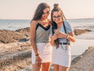 Улыбающиеся модные молодые женщины с помощью смартфона на пляже — стоковое фото