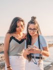 Sorrindo jovens mulheres na moda usando smartphone na praia — Fotografia de Stock