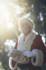 Вдумчивый пожилой человек в костюме Санта-Клауса, стоящий с подарком и колокольчиком в руках в перчатках на фоне природы — стоковое фото