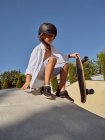 From below of happy boy wearing helmet holding skateboard sitting on ramp against blue sky — Stock Photo