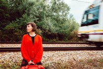 Stylische Frau im roten Pullover mit rotem Koffer, die bei der Schnellzugfahrt auf der Schiene hinter dem Rücken mitschaut — Stockfoto