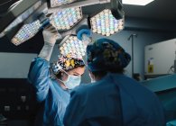 Серьезный молодой врач в защитной маске и кепке делает операцию с инструментами и медсестрой — стоковое фото