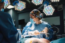 Серьезный молодой врач в защитной маске и кепке делает операцию с инструментами и медсестрой — стоковое фото