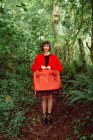 Donna in rosso con valigia rossa vintage in piedi nella foresta — Foto stock