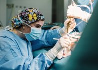 Vue latérale du jeune médecin sérieux en masque protecteur et capuchon faisant la chirurgie avec des instruments et infirmière de culture — Photo de stock