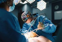 Ernsthafter junger Arzt in Schutzmaske und Mütze, der mit Instrumenten und Erntehelfer operiert — Stockfoto