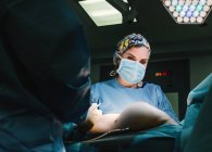 Снизу серьезный молодой врач в защитной маске и кепке делает операцию с инструментами и медсестрой — стоковое фото