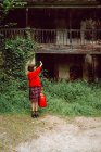 Mulher de vermelho com grande mala vermelha vintage em pé na frente da casa abandonada no campo — Fotografia de Stock