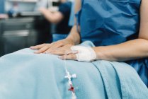 Crop female patient sitting with covered legs and intravenous fluid needle in hand before surgery in operating room — Stock Photo