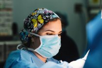 Side view of serious young doctor in protective mask and cap making surgery with instruments and crop nurse — Stock Photo