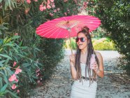 Весела струнка молода жінка в літньому вбранні і сонцезахисних окулярах з парасолькою, що п'є напій біля квітучих дерев — стокове фото