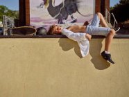 Vista lateral do menino de camisa deitado na borda da rampa em luz solar brilhante e refrigeração no parque de skate — Fotografia de Stock