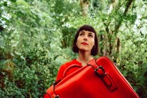 Жінка в червоному з великою червоною валізою, що йде в зеленому лісі — стокове фото