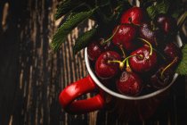 Вкусные аппетитные спелые вишни с листьями в красной чашке на темном деревянном столе — стоковое фото