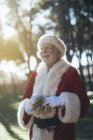 Homme âgé joyeux en costume du Père Noël debout avec présent et cloche dans les mains gantées sur fond de nature — Photo de stock