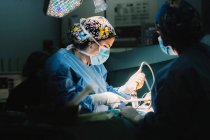 Side view of serious young doctor in protective mask and cap making surgery with instruments and crop nurse — Stock Photo