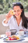 Woman sitting at table with served breakfast on open sunlit patio while holding and demonstrating donut and laughing with eyes closed on blurred background — Stock Photo