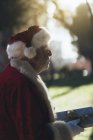 Homme âgé réfléchi en costume du Père Noël debout avec présent sur fond de nature — Photo de stock