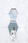 Estilo hombre afroamericano bien vestido que navega en la pierna móvil inclinándose en la pared geométrica en diseño futurista en el centro de la ciudad. - foto de stock