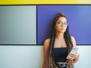 Pretty teenage girl with stylish dreadlocks using smartphone in colorful room — Stock Photo