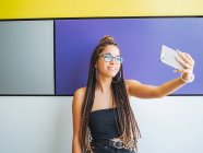 Pretty teenage girl with stylish dreadlocks taking selfie on smartphone in colorful room — Stock Photo
