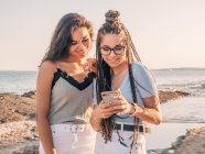 Щасливі щасливі випадкові жінки переглядають на смартфоні, стоячи на березі моря на заході сонця — стокове фото